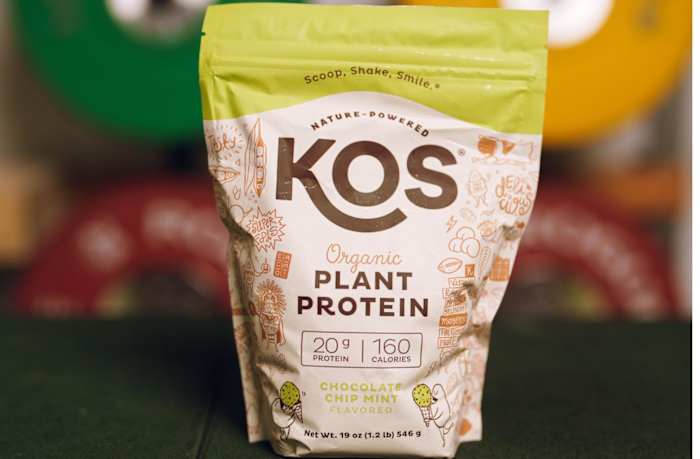 A bag of KOS Organic Plant Protein in Chocolate Chip Mint flavor and blurred weight plates in the background